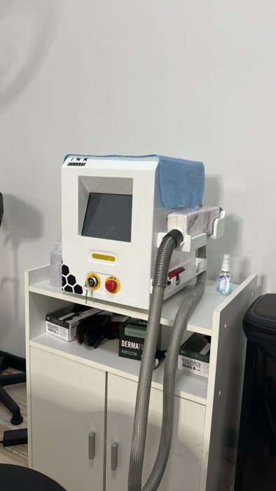 Una máquina láser médica o cosmética compacta, ideal para procedimientos de eliminación de tatuajes, se encuentra sobre un mueble con varios botones de control y una pantalla de visualización. Una manguera transparente se extiende desde el dispositivo. Debajo de ella, se ven cajas y una pequeña botella de gel desinfectante en el estante del mueble.