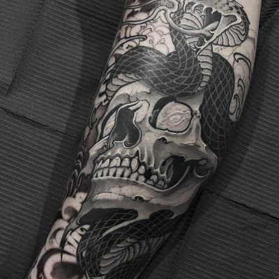 Un detallado tatuaje en negro y gris que cubre un antebrazo realizado por Carlos Benvenuto. El diseño presenta una calavera amenazante con múltiples serpientes enroscadas a su alrededor. El fondo incluye patrones ahumados y en espiral que realzan el tema oscuro y siniestro del tatuaje.