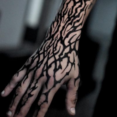 Primer plano de un brazo adornado con tatuajes abstractos de tinta negra que se asemejan a ramas o venas de árboles y que cubren toda la longitud desde el antebrazo hasta los dedos. Realizado en un reconocido estudio de Barcelona, el fondo oscuro y borroso resalta a la perfección el intrincado diseño del tatuaje.