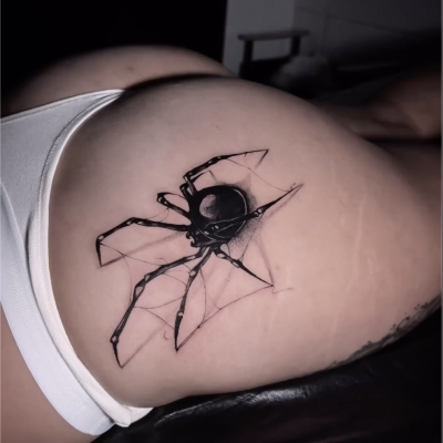 Una fotografía en primer plano de un gran tatuaje de una araña negra en la parte superior del muslo de una persona, realizado con intrincados detalles en Estudio en Barcelona. La persona lleva ropa interior blanca y las largas patas de la araña están estiradas con habilidad. El fondo está difuminado, lo que enfatiza el impresionante trabajo artístico del tatuaje.