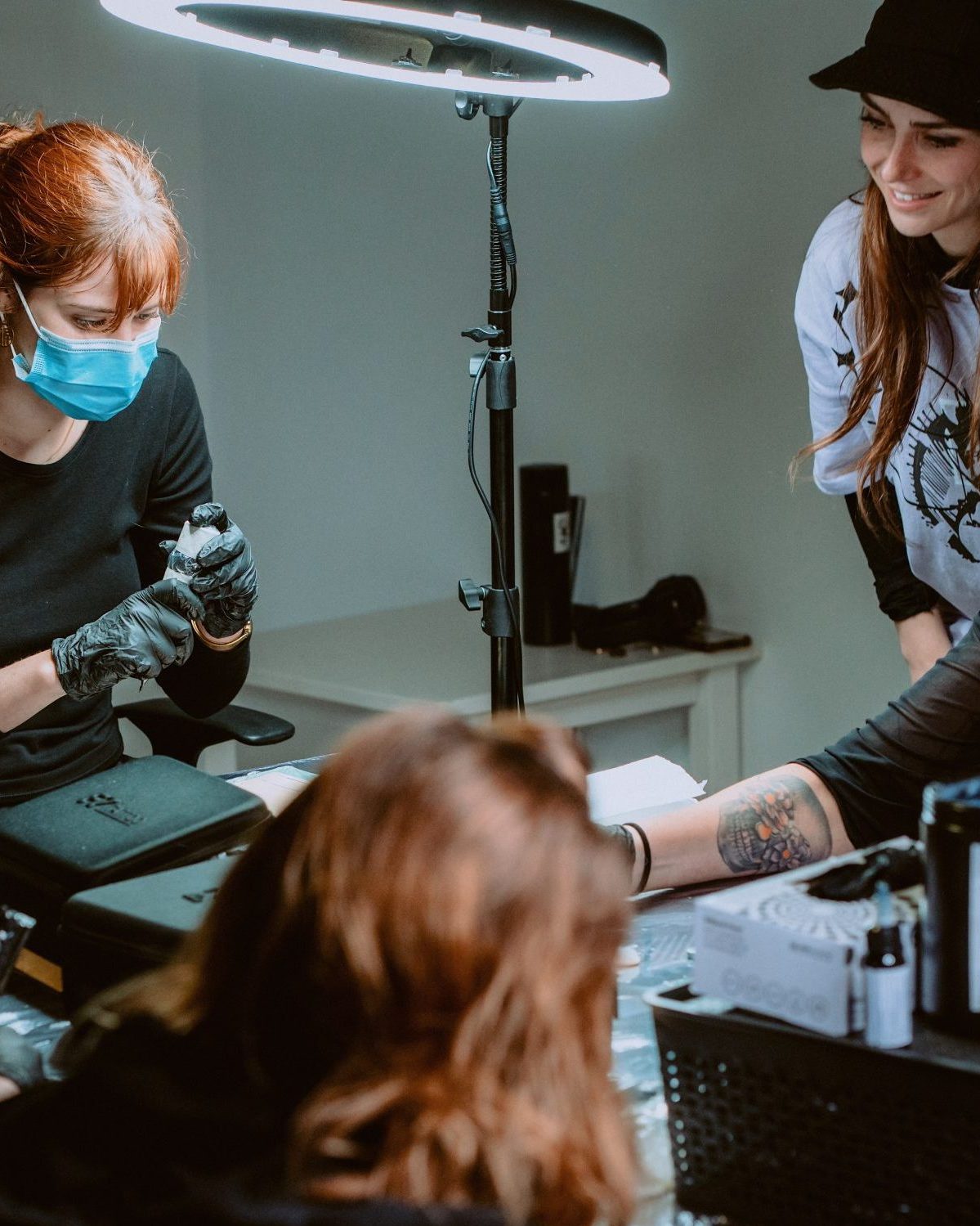 Cinco personas se encuentran en una sala bien iluminada reunidas alrededor de una mesa, tatuando o preparando el equipo para tatuar como parte de un curso de tatuajes. Una persona lleva una mascarilla y guantes, concentrada en su tarea. La escena es colaborativa y concentrada, con el equipo y las luces a la vista.