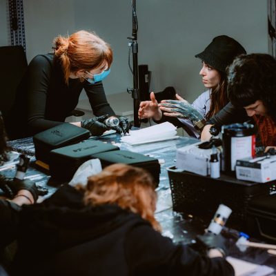 Un grupo de personas se reúne alrededor de una mesa, concentradas en sus tareas. La persona en el centro, que lleva una mascarilla, parece estar dando instrucciones a los demás. La mesa está llena de herramientas, cajas y equipos, lo que sugiere un entorno de taller para un curso de tatuajes.