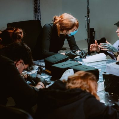 Un grupo de personas se reúne alrededor de una mesa, concentradas en sus tareas. La persona en el centro, que lleva una mascarilla, parece estar dando instrucciones a los demás. La mesa está llena de herramientas, cajas y equipos, lo que sugiere un entorno de taller para un curso de tatuajes.