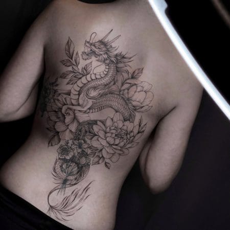 Una persona con un detallado tatuaje en tinta negra en la espalda. El tatuaje, que recuerda al vibrante arte que se suele ver en Barcelona, presenta un dragón entrelazado con grandes flores de peonía y un follaje intrincado que se extiende desde la parte superior de la espalda hasta la parte inferior. El fondo oscuro resalta la obra de arte.