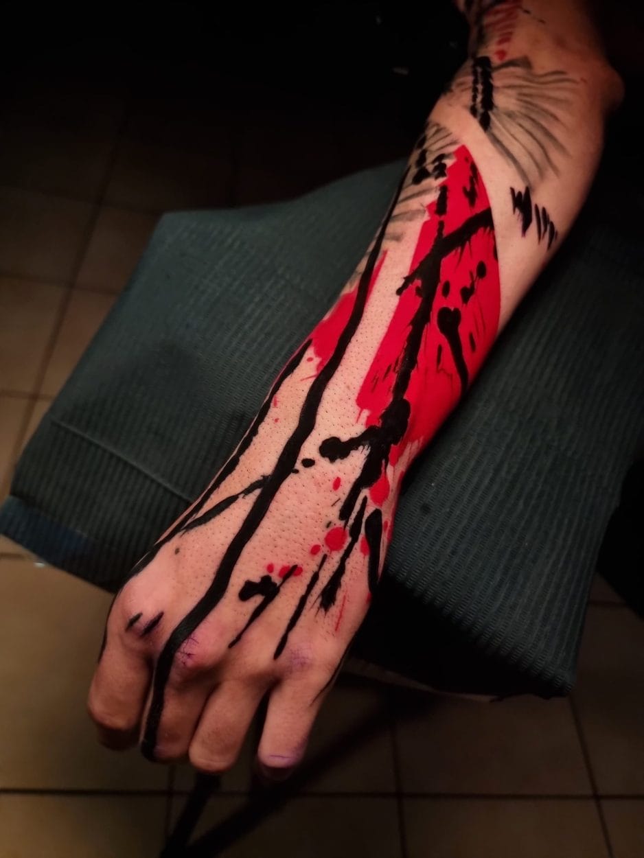 Un antebrazo con una mano extendida está tatuado con un diseño abstracto que presenta tinta roja y negra en un patrón salpicado. El brazo reposa sobre una superficie acolchada de color verde en Estudio Barcelona Tattoos. El fondo que lo rodea está tenuemente iluminado, lo que resalta el intrincado tatuaje.