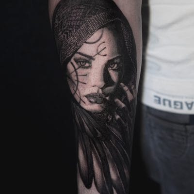 Primer plano de un tatuaje en el brazo de una persona que representa un retrato detallado y sombreado de una mujer con ojos intensos y que lleva una capucha. Tiene plumas y patrones intrincados alrededor de su rostro. El tatuaje está en tonos negros y grises, con la ropa de la persona visible en el fondo: un verdadero arte de los mejores tatuadores de Barcelona.