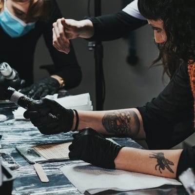 Dos personas, una de ellas con mascarilla, trabajan en una mesa con diversas herramientas y materiales. La persona en primer plano tiene tatuajes y utiliza una herramienta mientras lleva guantes negros. Las manos de otra persona se extienden sobre la mesa. El entorno sugiere un taller o un proyecto creativo relacionado con un curso de higiene sanitaria.