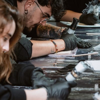 Las personas trabajan intensamente en tareas detalladas, utilizando herramientas o dispositivos portátiles y usando guantes negros. Una persona en el foco tiene tatuajes en el brazo y está inclinada hacia adelante, concentrada en su trabajo. El entorno se asemeja a un taller o estudio donde podría estar teniendo lugar un curso de higiene sanitaria.